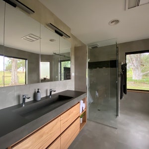 May include: A bathroom featuring a long vanity with a dark gray countertop, a rectangular integrated sink, and light wood cabinetry. Above the vanity is a large three-panel mirror. To the right is a glass-enclosed shower and a window.