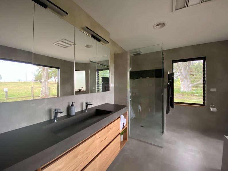 May include: A bathroom featuring a long vanity with a dark gray countertop, a rectangular integrated sink, and light wood cabinetry. Above the vanity is a large three-panel mirror. To the right is a glass-enclosed shower and a window.
