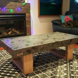 May include: A grey concrete coffee table with a speckled finish and a wooden base. The table is sitting on a black and white rug in front of a fireplace.
