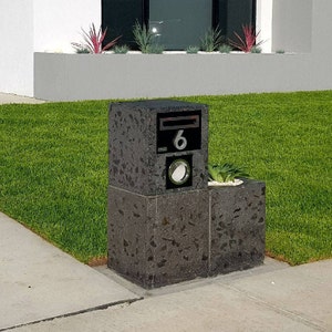 May include: A black mailbox with the number 6 on it, mounted on a concrete base with a small planter on top. The mailbox is surrounded by green grass.