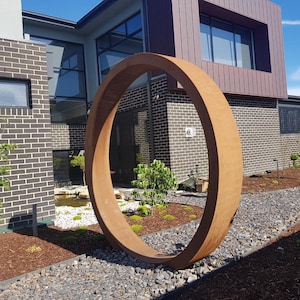May include: A large, rusty-colored metal ring sculpture in a modern garden setting. The ring is positioned in front of a brick house with large windows. The sculpture is surrounded by gravel and small trees.