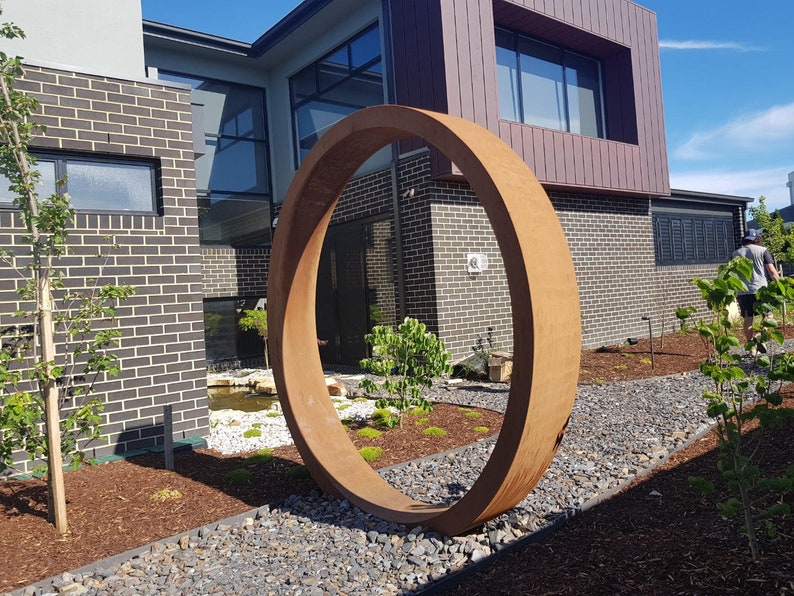 May include: A large, rusty-colored metal ring sculpture in a modern garden setting. The ring is positioned in front of a brick house with large windows. The sculpture is surrounded by gravel and small trees.