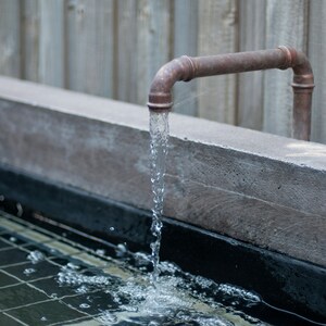 May include: A copper pipe spout with water flowing into a concrete basin with a black tiled bottom.