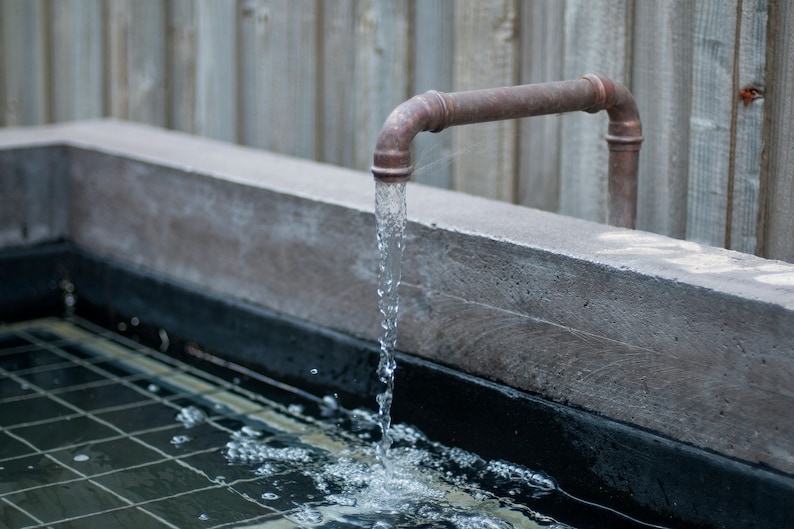 May include: A copper pipe spout with water flowing into a concrete basin with a black tiled bottom.
