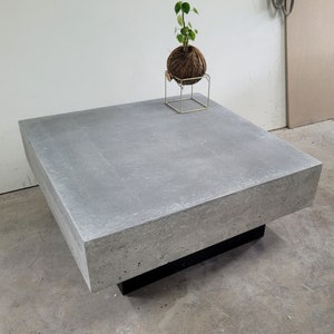 May include: A large, square, concrete coffee table with a black base. The table has a rough, textured surface and is made of a light gray concrete.