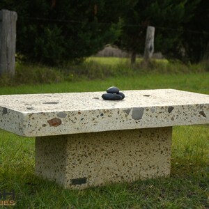May include: A large, rectangular concrete table with a light beige finish and a speckled surface. The table has a single, thick base and is sitting on a grassy lawn. Three small, black stones are stacked on top of the table.