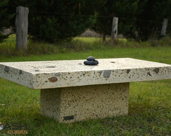 May include: A large, rectangular concrete table with a light beige finish and a speckled surface. The table has a single, thick base and is sitting on a grassy lawn. Three small, black stones are stacked on top of the table.