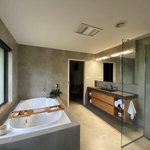 May include: A modern bathroom featuring a white oval soaking tub with a wooden tray, a floating wooden vanity with a dark countertop, dual sinks, and a glass-enclosed shower. The walls are covered in large gray tiles, and the floor has light tiles.