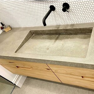 May include: A rectangular concrete sink basin integrated into a matching concrete countertop, featuring a matte black wall-mounted faucet and handle. The vanity below is constructed from light-toned wood with horizontal recessed drawer pulls.