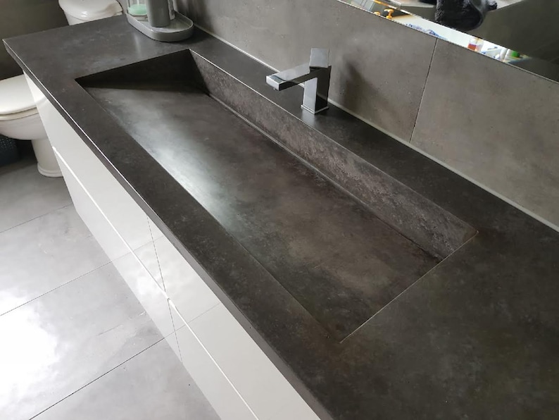 May include: A rectangular dark gray concrete bathroom sink integrated into a matching countertop. The basin features a flat, sloped bottom and straight edges. A polished chrome single-handle faucet is mounted on the rear deck of the sink.