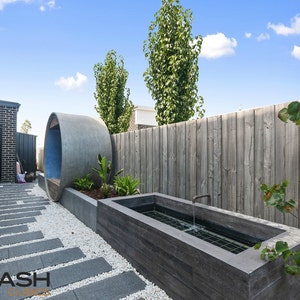 May include: A modern backyard with a concrete water feature, a stone walkway, and a large concrete pipe sculpture. The backyard is surrounded by a wooden fence and green trees.