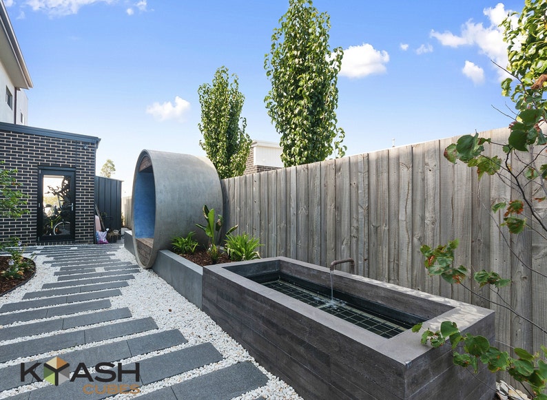 May include: A modern backyard with a concrete water feature, a stone walkway, and a large concrete pipe sculpture. The backyard is surrounded by a wooden fence and green trees.