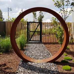 May include: A large, rusty-colored metal ring stands in a garden setting. The ring is positioned in front of a wooden fence and a black gate. The ring is large enough to frame the gate and the fence. The ground is covered in brown mulch and small gray stones.