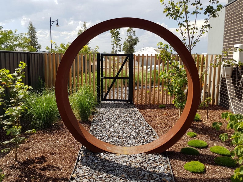 May include: A large, rusty-colored metal ring stands in a garden setting. The ring is positioned in front of a wooden fence and a black gate. The ring is large enough to frame the gate and the fence. The ground is covered in brown mulch and small gray stones.
