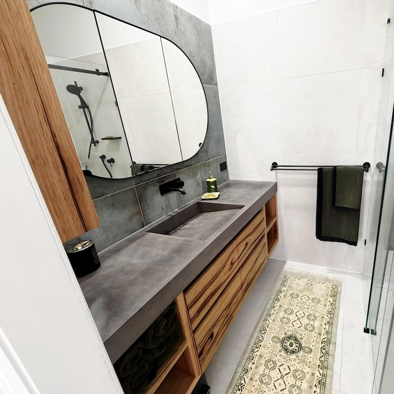 May include: A bathroom vanity featuring a dark gray concrete countertop with an integrated rectangular sink, a wooden cabinet base with open shelving, a large arched mirror, and a black wall-mounted faucet, set against gray tiled walls.