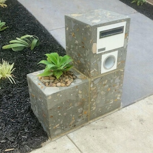 May include: A concrete mailbox with a white door and a silver mail slot. The mailbox is attached to a concrete planter with a green plant inside. The planter and mailbox are both made of a textured concrete with small stones embedded in the surface.