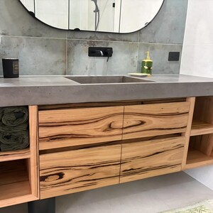 May include: A bathroom vanity featuring a gray concrete countertop with an integrated sink, four light wood drawers with prominent dark grain patterns, and open side shelving. A black faucet is mounted on the gray tiled wall above the sink.