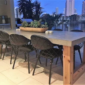May include: A concrete dining table with a wooden base and six black metal chairs. The table is set with coasters and the chairs are arranged around the table. The table is on a patio overlooking a body of water with buildings in the background.