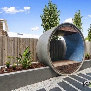 May include: A concrete pipe shaped into a half-circle creates a unique outdoor seating area. The pipe is painted blue on the inside and has a wooden bench seat. The seating area is surrounded by gravel and a concrete planter with green plants.