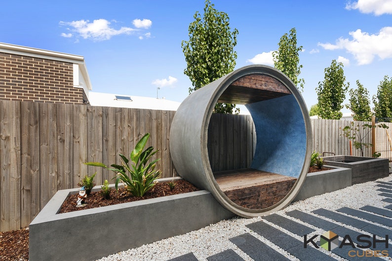 May include: A concrete pipe shaped into a half-circle creates a unique outdoor seating area. The pipe is painted blue on the inside and has a wooden bench seat. The seating area is surrounded by gravel and a concrete planter with green plants.