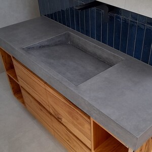 May include: A bathroom vanity featuring a rectangular gray concrete sink basin integrated into a smooth concrete countertop. The base is constructed from light brown wood with open shelving and drawers. A matte black faucet is mounted above the sink.