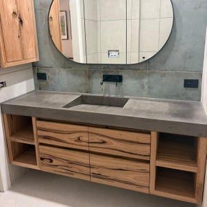 May include: A bathroom vanity featuring a gray concrete countertop with an integrated rectangular sink, mounted above a wooden cabinet with two central drawers and open side shelving, paired with a large oval mirror and matte black faucet.