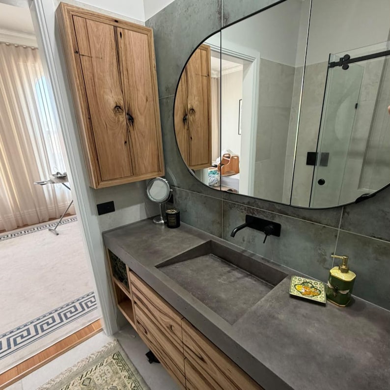 May include: A bathroom vanity featuring a dark gray concrete countertop with an integrated rectangular sink, a wall-mounted black faucet, and light-toned wood cabinetry. Above the sink is a large oval mirror and a matching wood wall cabinet.
