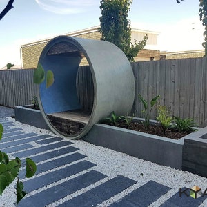 May include: A concrete pipe with a wooden bench inside, used as a unique outdoor seating area. The pipe is surrounded by white gravel and a small garden with plants. A stone path leads to the seating area.