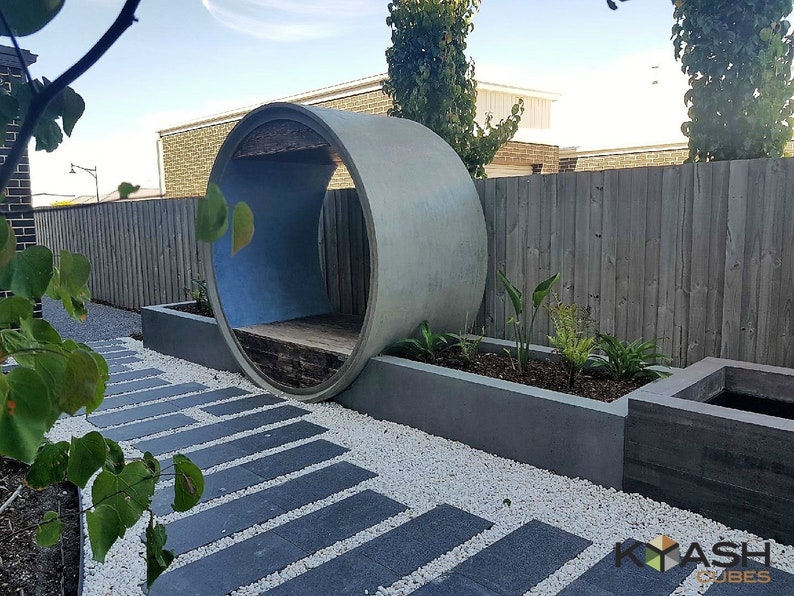 May include: A concrete pipe with a wooden bench inside, used as a unique outdoor seating area. The pipe is surrounded by white gravel and a small garden with plants. A stone path leads to the seating area.
