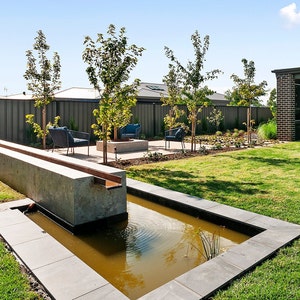 May include: A modern backyard with a concrete water feature, a small pond, and a seating area with chairs and a fire pit. The backyard is surrounded by a fence and lush green grass.