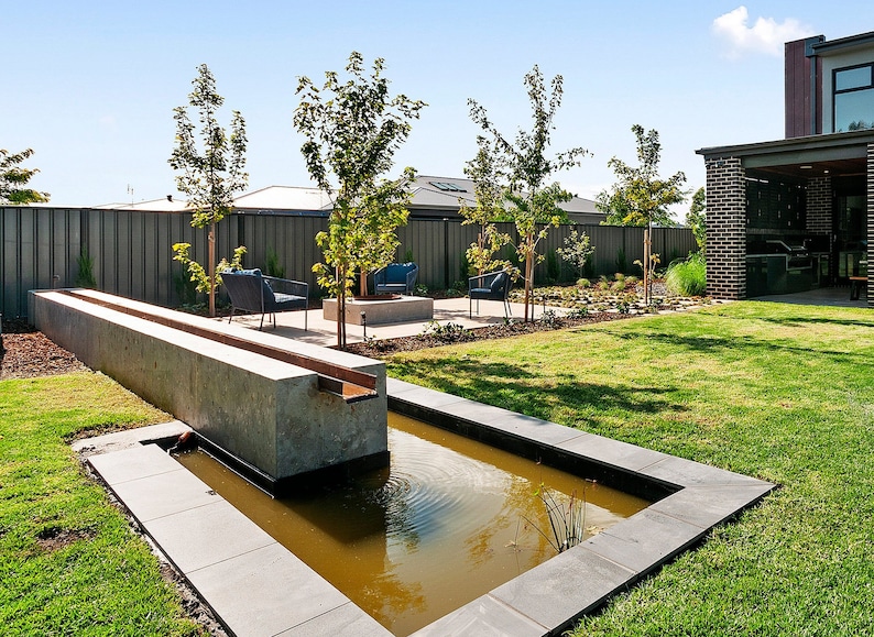 May include: A modern backyard with a concrete water feature, a small pond, and a seating area with chairs and a fire pit. The backyard is surrounded by a fence and lush green grass.