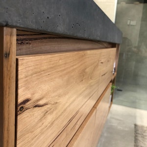 May include: A bathroom vanity cabinet featuring light brown natural wood grain drawer fronts with visible knots and dark streaks, topped with a thick, dark gray concrete countertop that has a textured, porous surface finish.