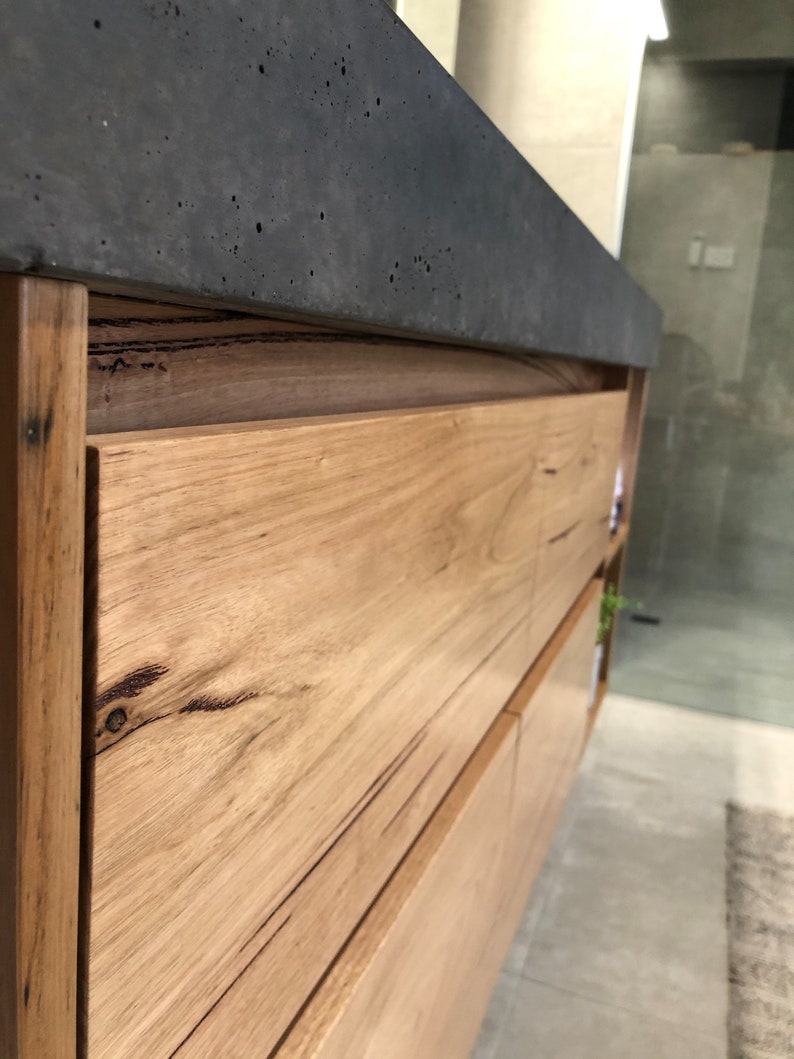 May include: A bathroom vanity cabinet featuring light brown natural wood grain drawer fronts with visible knots and dark streaks, topped with a thick, dark gray concrete countertop that has a textured, porous surface finish.
