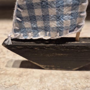 Puede incluir: Un peque&ntilde;o velero de madera con una vela a cuadros azul y blanco. La vela es de tela y est&aacute; cosida con hilo blanco. El casco del barco es de color marr&oacute;n oscuro.
