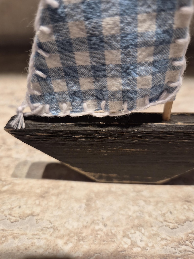 Puede incluir: Un peque&ntilde;o velero de madera con una vela a cuadros azul y blanco. La vela es de tela y est&aacute; cosida con hilo blanco. El casco del barco es de color marr&oacute;n oscuro.
