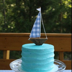 Puede incluir: Un pastel peque&ntilde;o y redondo con glaseado azul y un velero decorativo. El velero tiene una vela a cuadros azul y blanco y una peque&ntilde;a bandera blanca. El pastel est&aacute; sobre un plato de vidrio transparente.