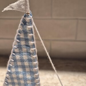 Puede incluir: Un velero en miniatura hecho a mano con una vela de cuadros azules y blancos y una peque&ntilde;a bandera blanca. La vela est&aacute; cosida con hilo blanco, y una cuerda blanca se extiende desde la parte superior. El barco es de madera.