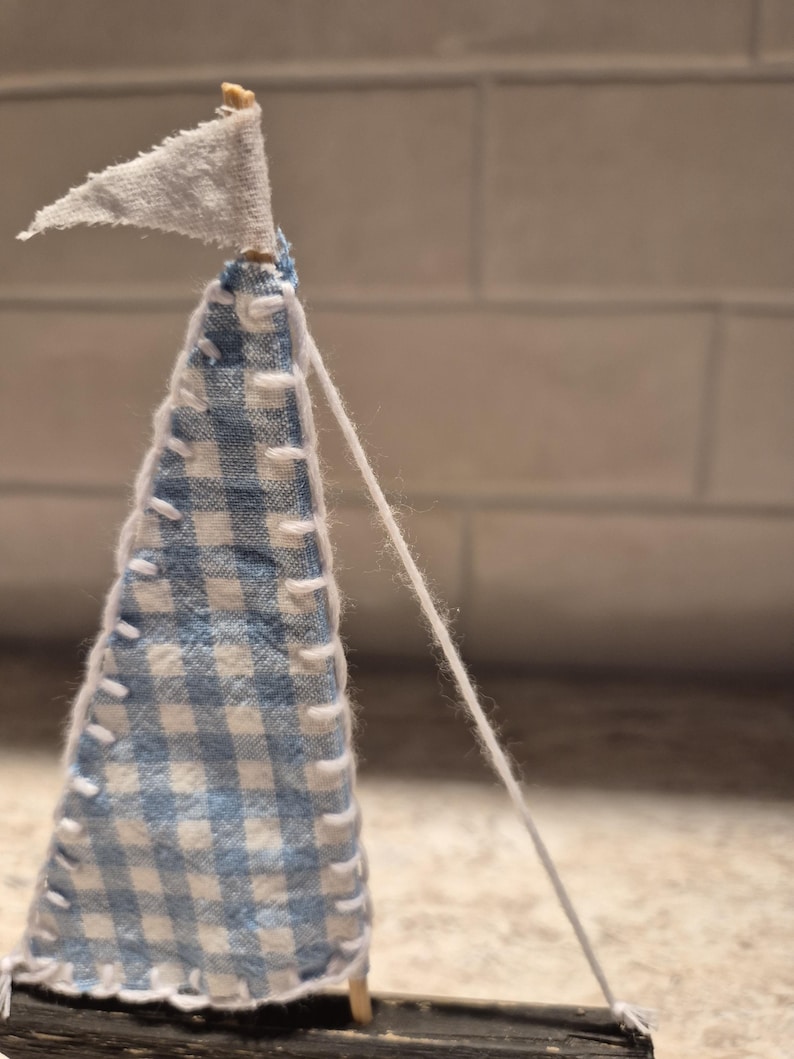 Puede incluir: Un velero en miniatura hecho a mano con una vela de cuadros azules y blancos y una peque&ntilde;a bandera blanca. La vela est&aacute; cosida con hilo blanco, y una cuerda blanca se extiende desde la parte superior. El barco es de madera.