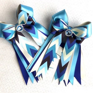 May include: Two blue ribbon bows with chevron patterned tails. Each bow features a central jewel and is made of satin ribbon in shades of blue, white, and black. The bows are likely for equestrian use or as decorative accessories.
