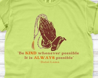 Può includere: Una maglietta verde chiaro con un'illustrazione marrone di mani in preghiera che tengono una collana di perline. Il testo "Be KIND whenever possible It is ALWAYS possible" è stampato sotto l'illustrazione, con il testo " - Dalai Lama" stampato sotto.