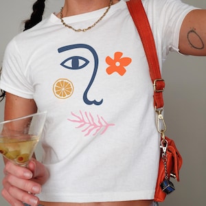 May include: White t-shirt with a graphic design of a stylized face with a blue eye, orange citrus fruit, pink coral, and a red flower.