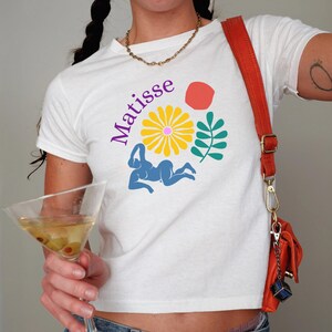 May include: White cropped t-shirt with a purple "Matisse" text, a blue female figure, a yellow flower, green leaves, and a red circle graphic.