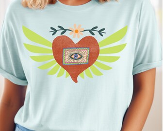 Könnte beinhalten: Ein hellblaues T-Shirt mit einem Grafikdruck eines roten Herzens mit Flügeln und einem Auge darin. Das Herz hat ein braunes, orangefarbenes und gelbes Design. Die Flügel sind grün.