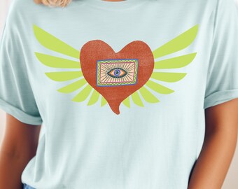 Könnte beinhalten: Ein hellblaues T-Shirt mit einem roten Herz mit Flügeln. Das Herz enthält ein blaues Auge mit bunten Details.