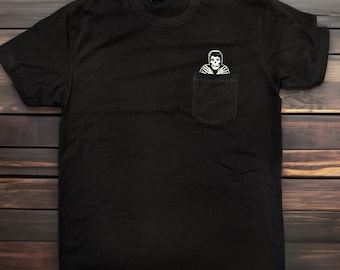 May include: Black t-shirt with a pocket featuring a white graphic of a skeleton in a hooded sweatshirt.