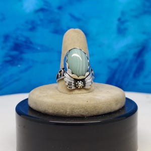 May include: A silver ring with a green and white oval stone set in a decorative silver setting. The ring is on a display stand.