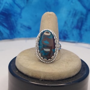 May include: Oval-shaped silver ring featuring a large, polished gemstone with teal and brown hues. The gemstone is set in a detailed, beaded bezel. The ring is displayed on a neutral-toned ring holder against a blue backdrop.