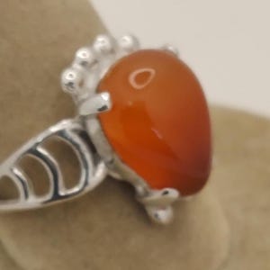 May include: A silver ring featuring a large, teardrop-shaped, orange-red gemstone. The ring has a decorative silver setting with a detailed design on the band and around the gemstone. The gemstone is smooth and polished.