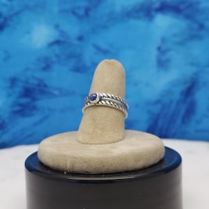 May include: A silver ring with a blue gemstone set in the center. The ring is on a display stand.