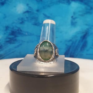 May include: A silver ring with a green agate stone set in a bezel setting. The ring has a textured band and is on a display stand.